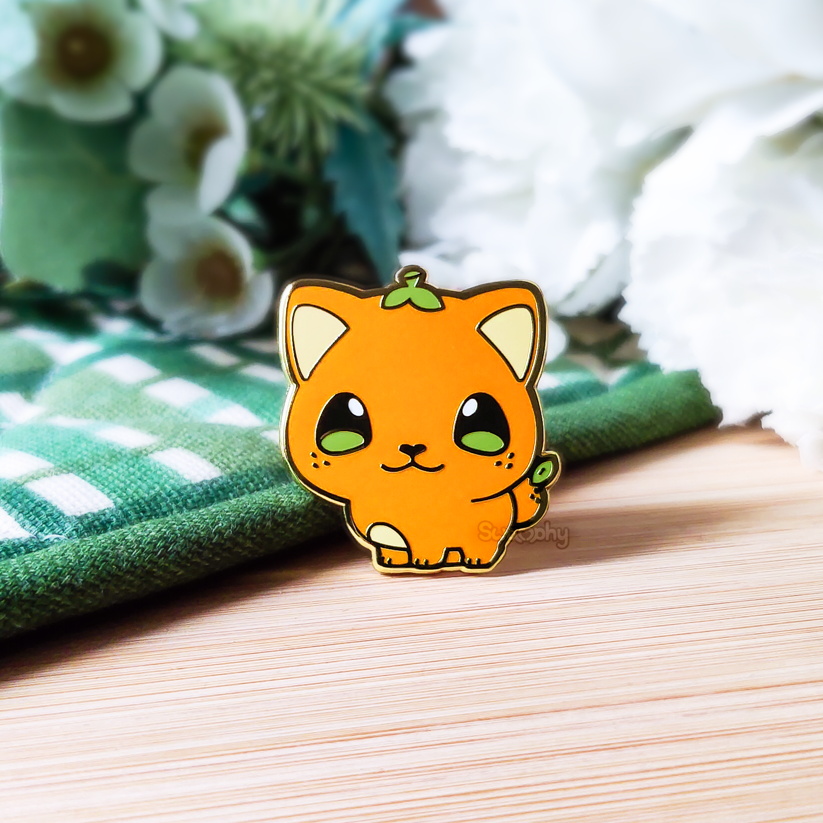 Photo of a hard enamel pin of an orange-themed bobtail cat. It is a vibrant orange with a green stem on top of its head, green eyes, and an orange as their tail with a leaf on top. It has three freckles on each cheek and tail, simulating the appearance of an orange's skin.