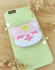 Photo of a PVC phone grip of a cute, pink Shiba Inu with a sakura mochi leaf on top of his head. He's sticking out his tongue with his eyes closed, smiling sweetly and cheekily at the viewer. The colors are vibrant, light, and pastel, and the phone grip is attached to a pastel green iPhone 6S case against a wooden background with dried flowers at the corners of the photo.